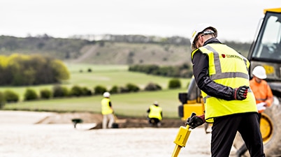 1st Line Defence operative conducting a UXO Search & Clear operation on a construction site in the UK with heavy machinery and fields in the background